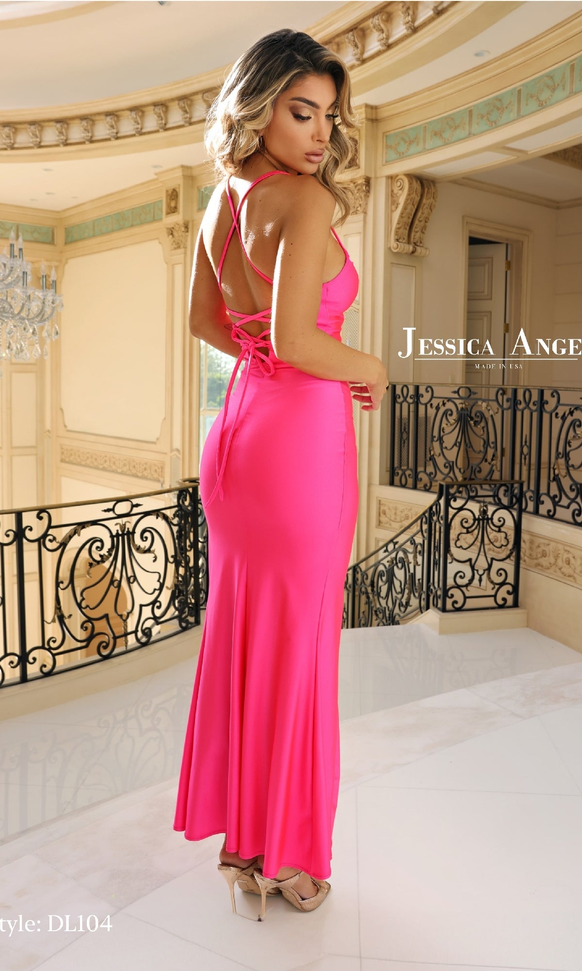 A woman wearing the Jessica Angel DL104 Lace-Up Long Prom Dress in bright pink stands indoors on a marble floor before ornate railings and beige walls.