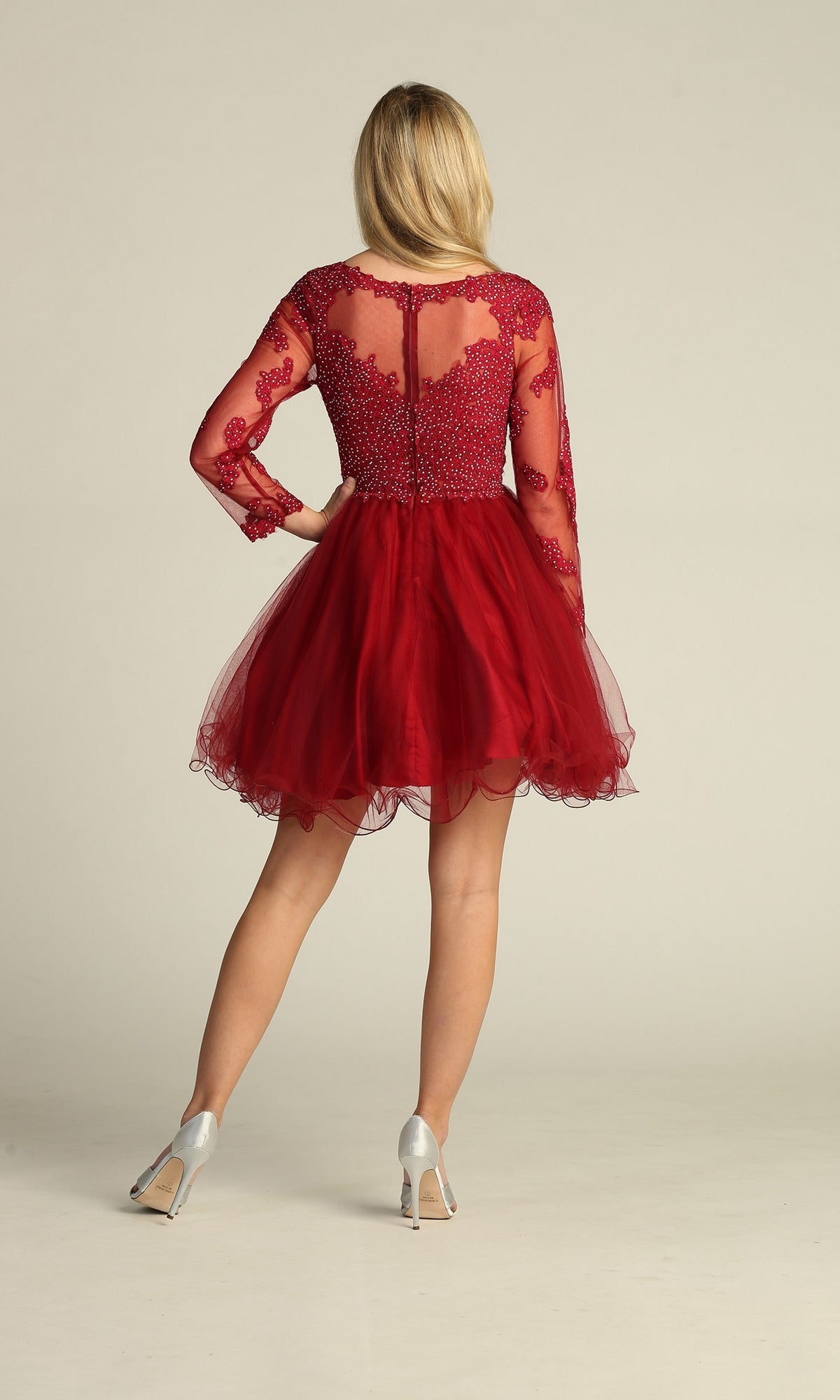 A woman faces away, wearing the Beaded Long Sleeve Short Homecoming Dress DM4003 with a sheer, beaded bodice and long sleeves, paired with silver high heels against a plain background.