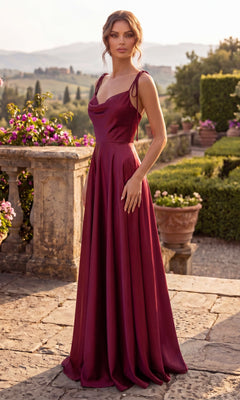 A woman wearing the Shoulder-Tie Long A-Line Prom Dress: Chicas DM4018 in burgundy stands on a stone terrace with greenery, flowers, and hills in the background.