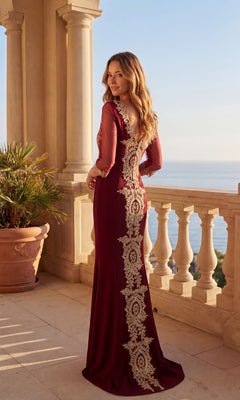 A woman in the Long Formal Dress: Chicas DM4020—burgundy with gold embroidery and a v-neckline—stands on a stone balcony overlooking the ocean, with a potted plant nearby.