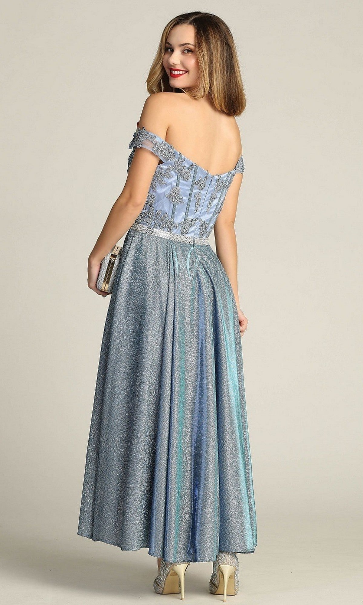 A woman stands smiling over her shoulder, wearing the Chicas DM4027 Tea-Length Formal Dress—a light blue off-the-shoulder style featuring sparkly fabric and an elegant embroidered bodice—her look perfectly complemented by the clutch bag in her hand.