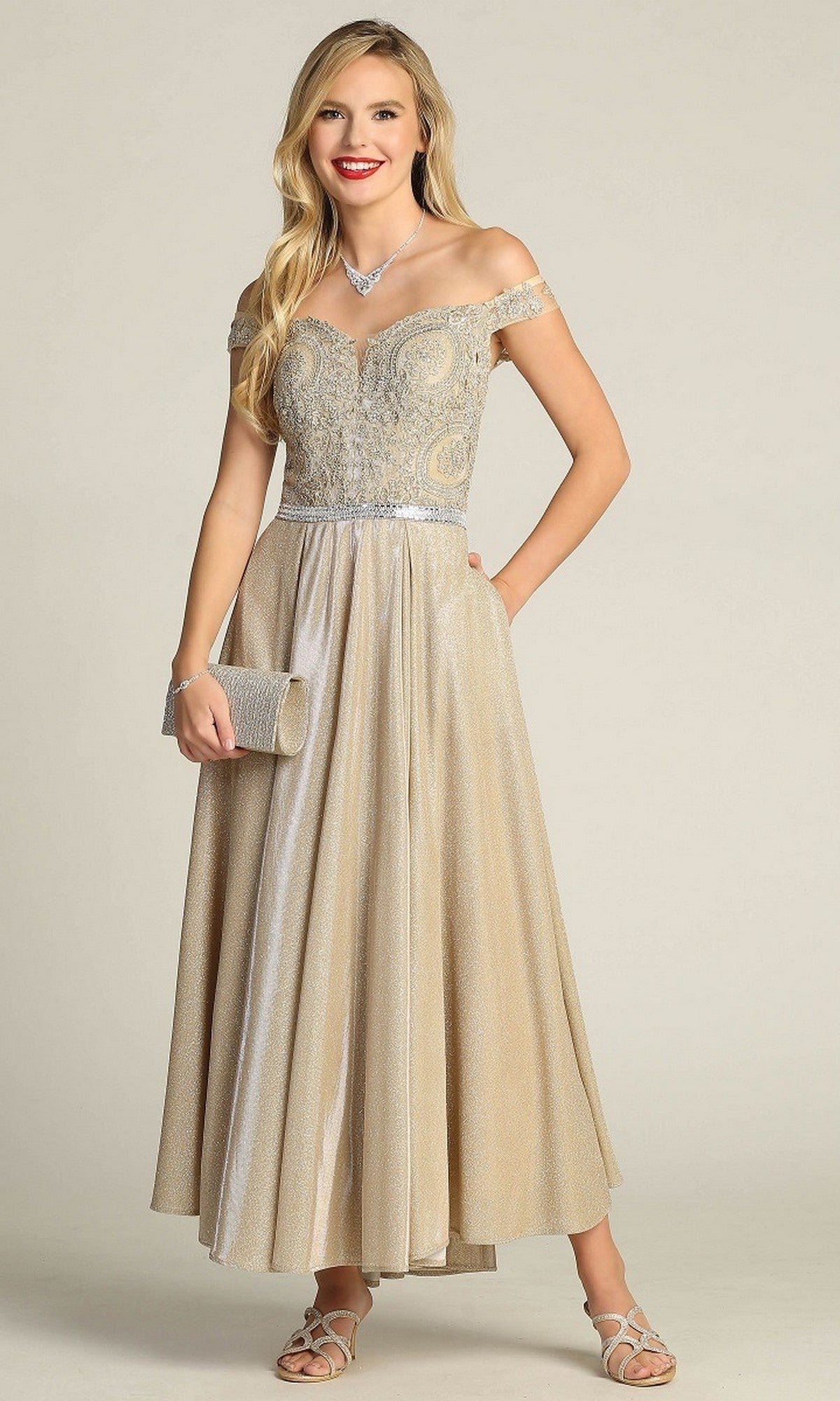 A woman in the Tea-Length Formal Dress: Chicas DM4027 in champagne gold, featuring a beautifully embroidered off-the-shoulder bodice, holds a matching clutch and accessorizes with sandals and a necklace while standing against a plain background.