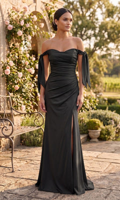 A woman wearing an elegant off-the-shoulder, black Long Prom Dress: Chicas DM8010 with a thigh-high slit stands outdoors near a stone bench and blooming flowers.