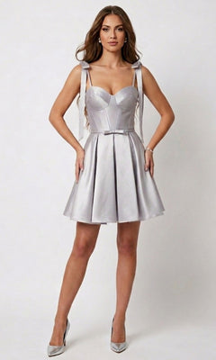 A woman wearing the Short Homecoming Dress: Chicas DM8133 in silver—a sleeveless, fit-and-flare satin dress with a corset bodice and belted waist—paired with silver high heels, stands against a plain light background.