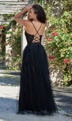 A woman stands outdoors by a wall of blooming pink flowers, wearing the Velvi Lace-Bodice Long Ruffled Prom Dress Dahlia featuring a lace bodice, crisscross back straps, and a flowing black A-line skirt.