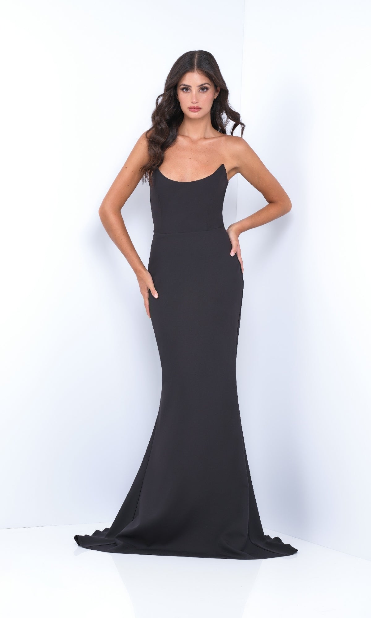 A woman poses against a white background wearing the Strapless Classic Black Prom Dress: Daris by Velvi—a floor-length black gown with a slight train.