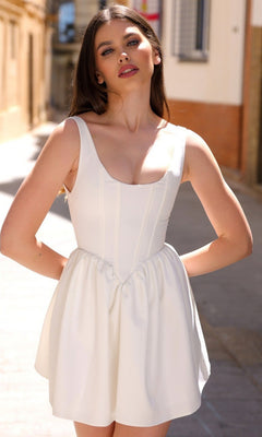 A woman stands outdoors on a sunlit street, wearing the Velvi A-Line Mini Graduation Party Dress Darling—a sleeveless, white fitted cocktail dress with a flared skirt, ideal as a short semi-formal dress for special occasions.
