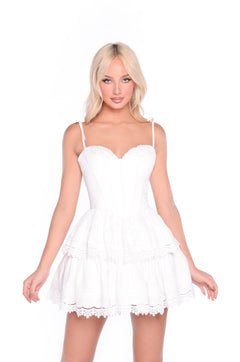 A woman with long blonde hair models the Delilah White Short Corset Graduation Dress, which features a corset bodice and lace-trimmed tiered skirt, standing against a plain white background.