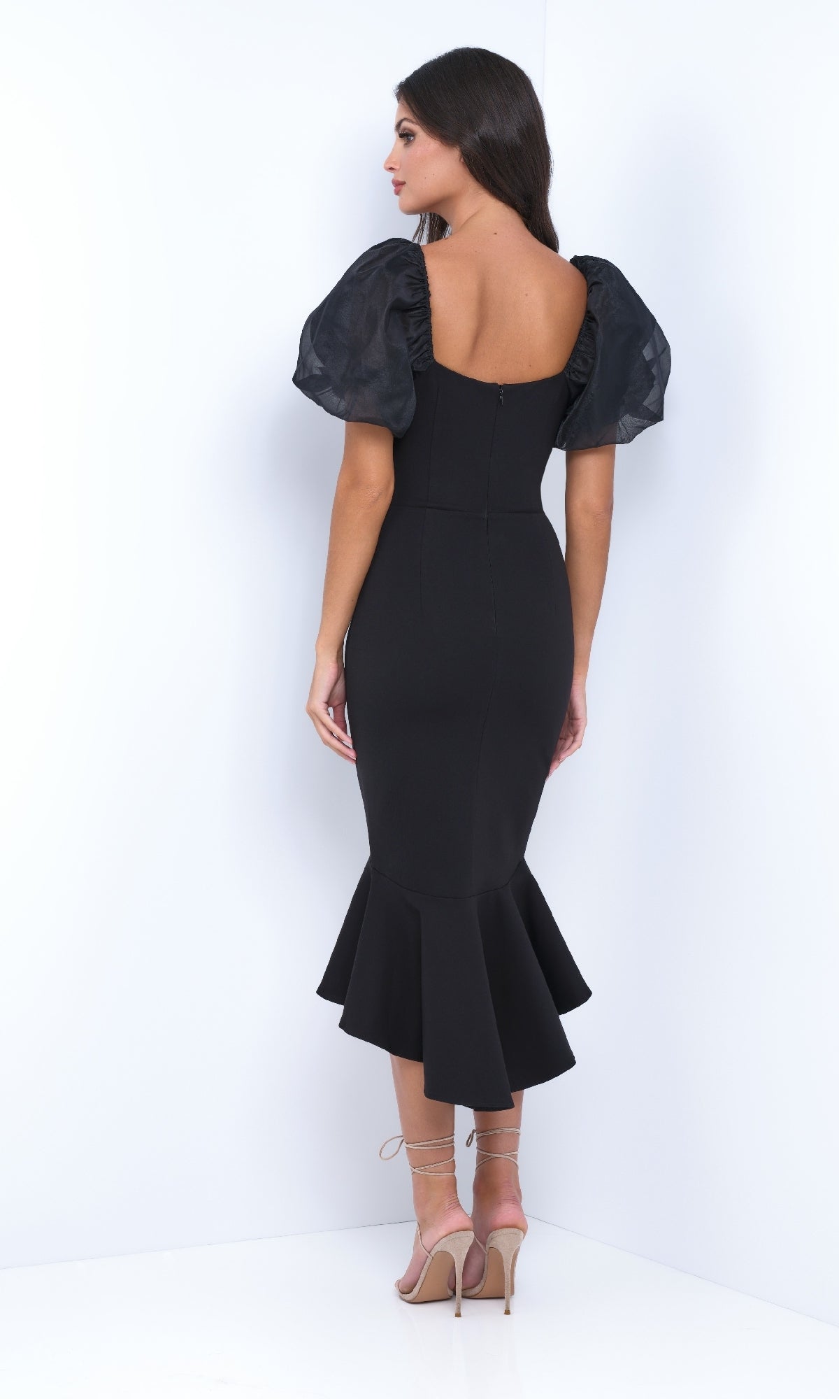 A woman stands in a corner wearing the Velvi Short Sleeve Midi Semi-Formal Dress Diana—fitted with puffed sleeves and a flared hem—styled with strappy nude heels, ideal for your next rehearsal dinner or evening event.