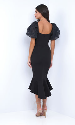 A woman stands in a corner wearing the Velvi Short Sleeve Midi Semi-Formal Dress Diana—fitted with puffed sleeves and a flared hem—styled with strappy nude heels, ideal for your next rehearsal dinner or evening event.