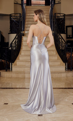 Wearing the Cowl-Neck Corset Pleated Satin Long Prom Dress E1042, a woman stands at the base of a staircase, her back to the camera as silver satin and crisscross straps catch the light.