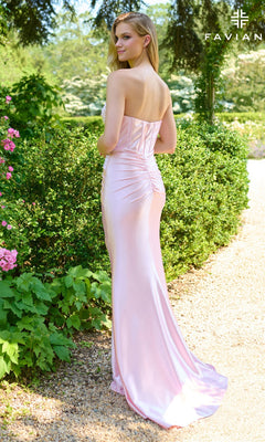 Wearing the Faviana E11422 long prom dress, a woman stands on a garden path among greenery and pink flowers, her strapless satin corset gown flowing as she faces away from the camera.