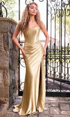 A woman standing by an iron gate in a gold Faviana E11632 long prom dress with a strapless corset bodice, floor-length silhouette, and high side slit.