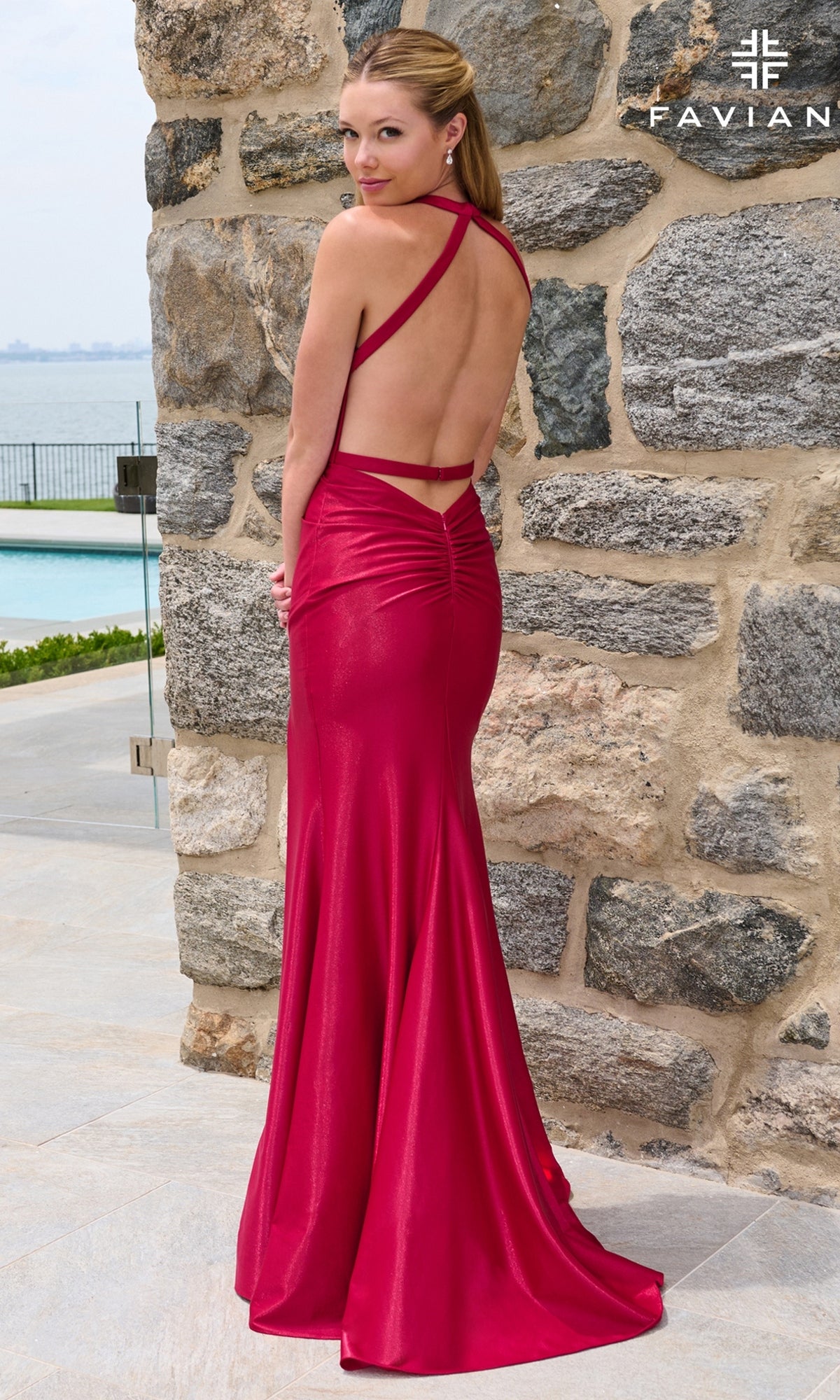 A woman standing by a stone wall wearing a floor-length red Faviana E11661 prom dress with an open back and crisscross straps.