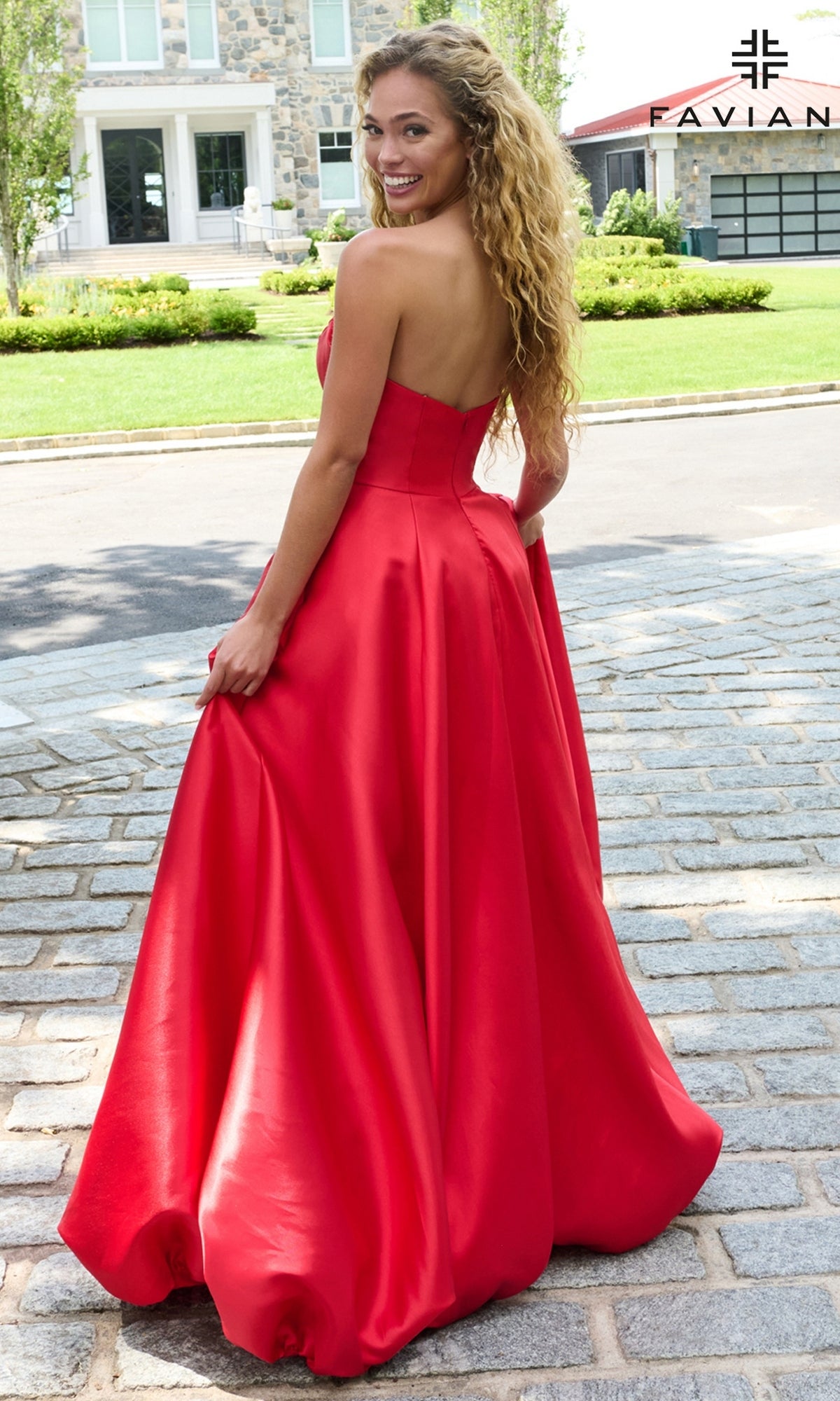 A woman in a strapless, floor-length red Faviana E11670 prom dress stands on a cobblestone path, looking over her shoulder.