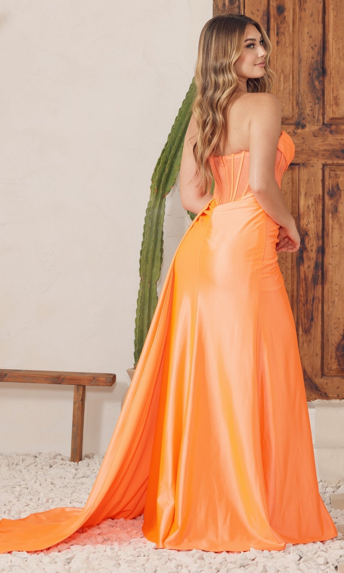 A woman in the Side Drape Long Sheer-Corset Prom Dress E1237, a strapless, floor-length orange gown with a long train, stands indoors near a cactus and wooden door, facing slightly away from the camera.
