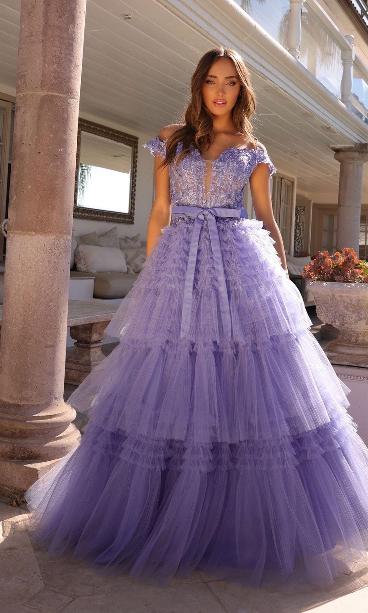 A woman stands outdoors beside a stone column in the Nox Anabel Designer Ruffled Prom Ball Gown E1293, a floor-length lavender tulle dress with beaded embroidered lace and a deep V-neck.