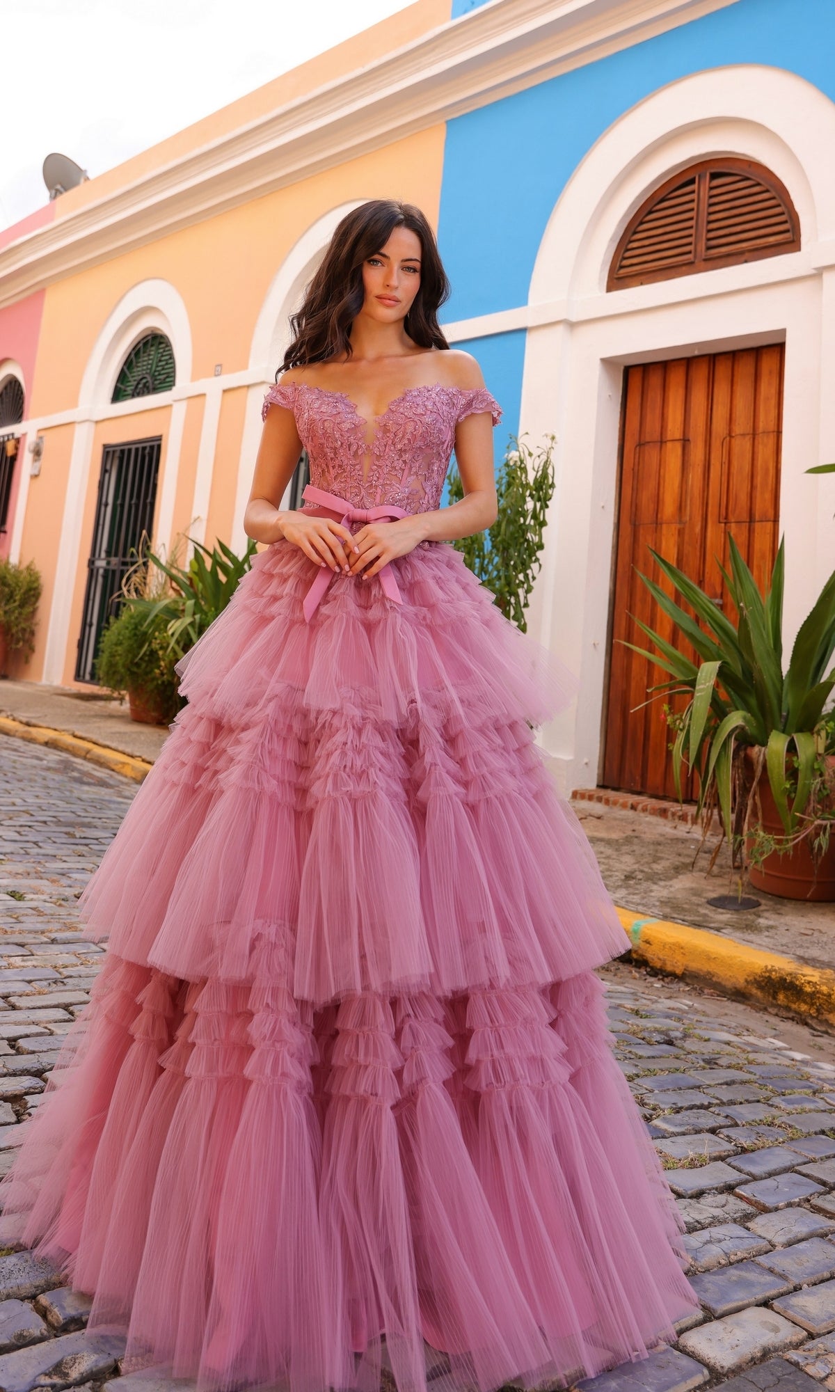 A woman stands on a cobblestone street wearing the Nox Anabel Designer Ruffled Prom Ball Gown E1293, layered with pink tulle, as colorful buildings and potted plants provide a charming backdrop.