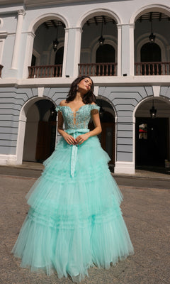 A woman in the Nox Anabel Designer Ruffled Prom Ball Gown E1293, featuring light blue off-the-shoulder beaded lace, stands before a white building with arched balconies.