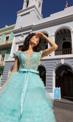 A woman in the Nox Anabel Designer Ruffled Prom Ball Gown E1293 stands outdoors, shielding her eyes from the sun in front of a white and pastel-colored building.