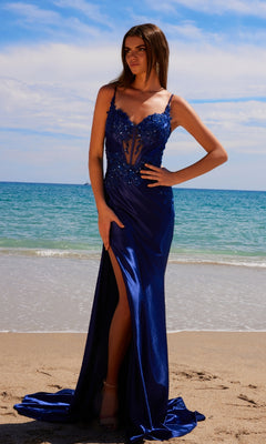 A woman in the Nox Anabel E1488 Long Corset Prom Dress with Train, featuring a thigh-high slit, stands on a sandy beach with the ocean and sky as a stunning backdrop.
