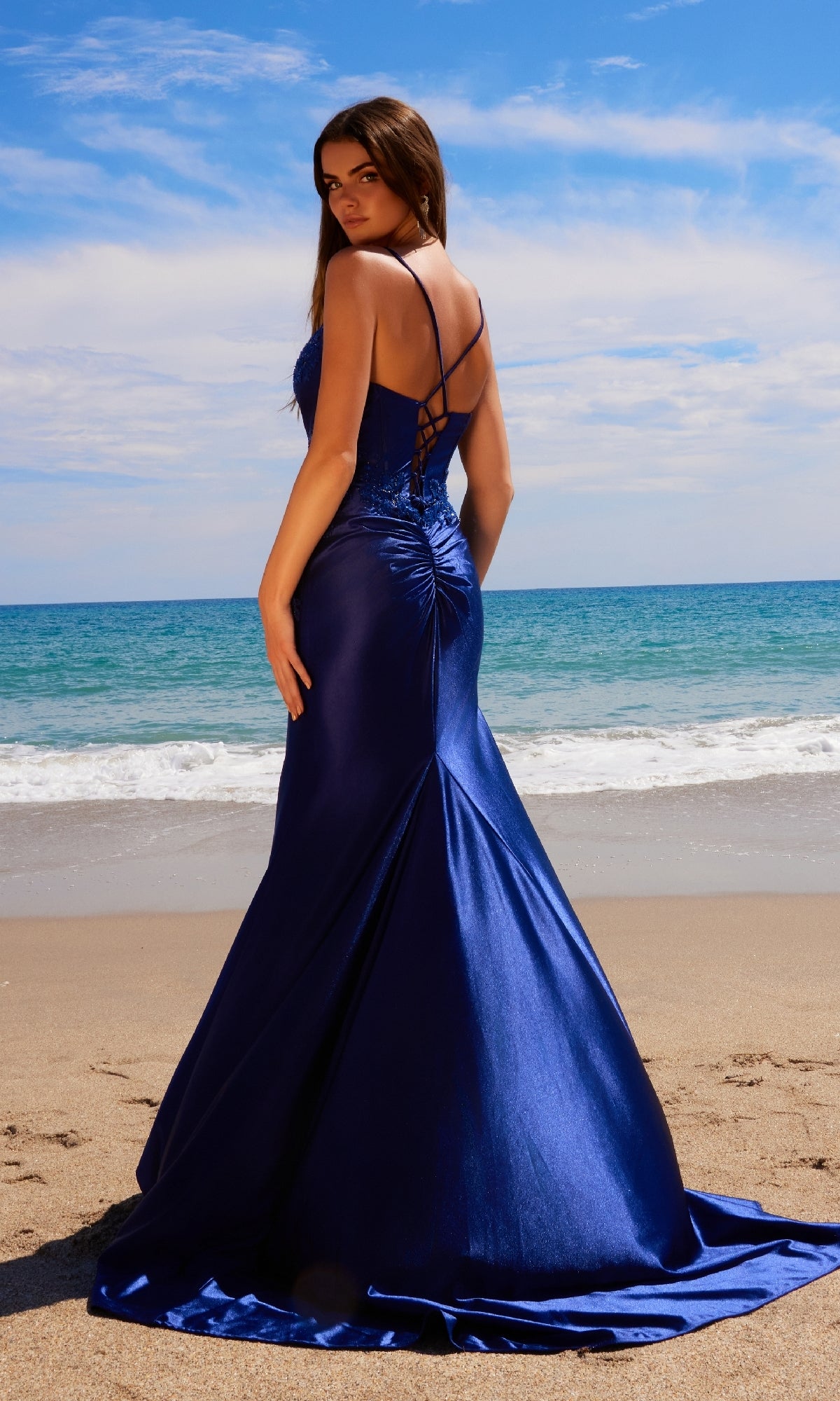 Woman in a fitted, floor-length Nox Anabel E1488 long corset prom dress with train and lace-up back stands on a sandy beach facing the ocean beneath a partly cloudy sky.