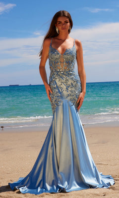 A woman stands on a sandy beach with the ocean and sky behind her, wearing the Nox Anabel E1532—a light blue, fitted, floor-length mermaid prom dress with beaded embroidery and floral embellishments.