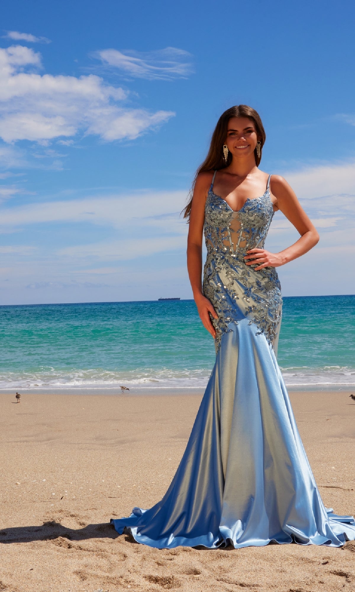A woman wearing the Nox Anabel E1532 Long Mermaid Prom Dress, with beaded embroidery in light blue, stands on a sandy beach against a beautiful ocean and sky backdrop.