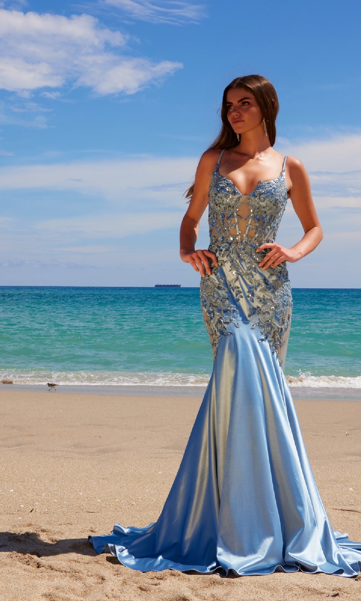 A woman wearing the Nox Anabel E1532 Long Mermaid Prom Dress in light blue with beaded embroidery stands on a sandy beach, the ocean and blue sky behind her.