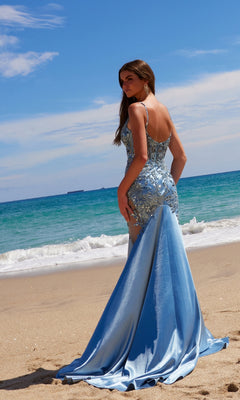 A woman in the Nox Anabel E1532 Long Mermaid Prom Dress, featuring light blue beaded embroidery, stands on a sandy beach looking out at the ocean beneath a partly cloudy sky.