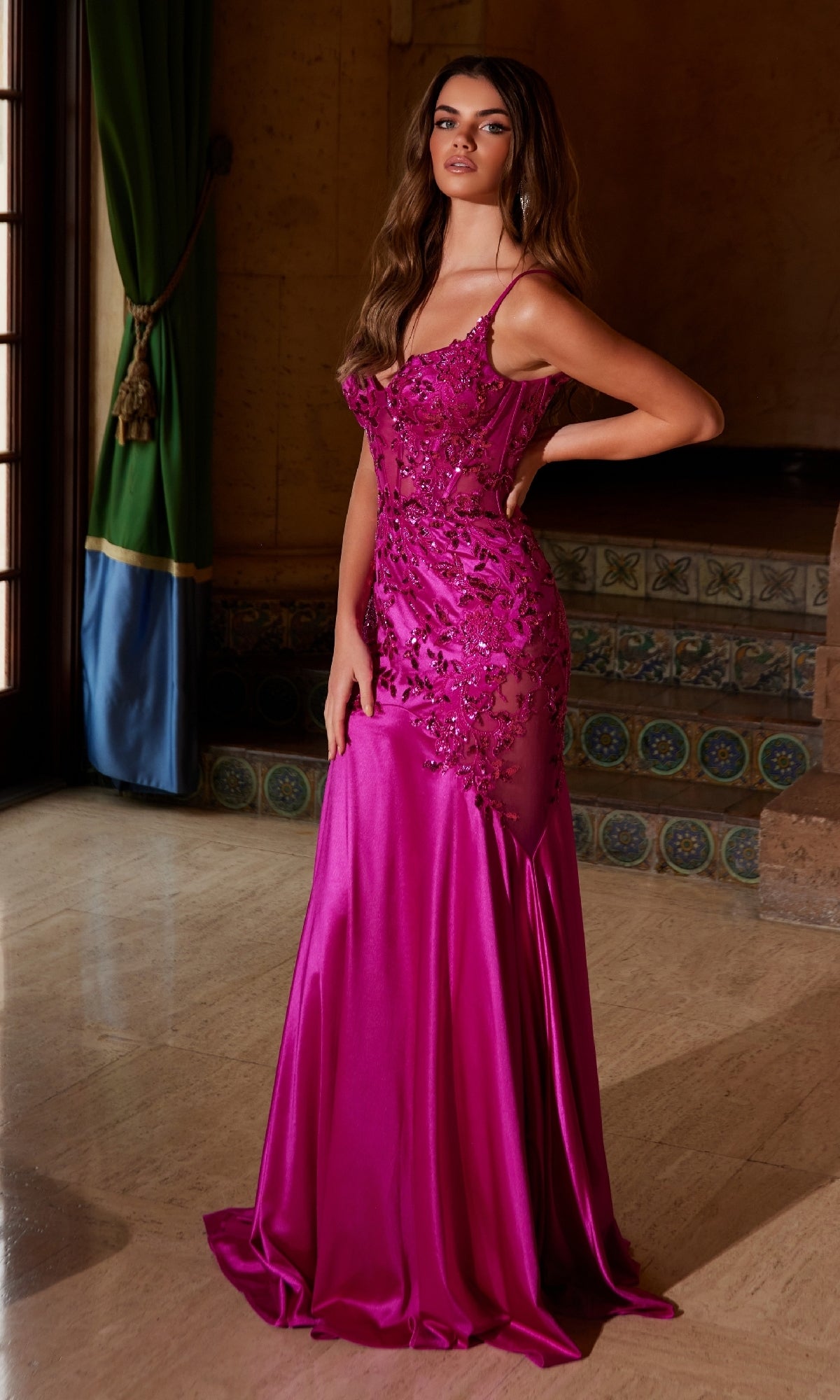 A woman poses indoors with one hand on her hip, wearing the Nox Anabel E1532— a floor-length magenta satin mermaid prom dress featuring floral embellishments, beaded embroidery, and sheer panels.