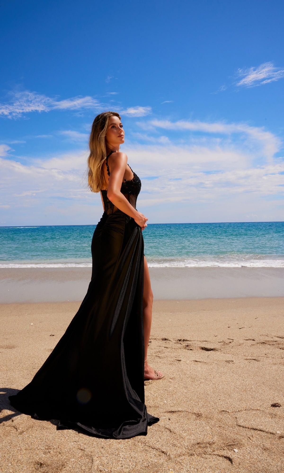 Wearing the Sheer-Waist Long Prom Dress: Nox Anabel E1711, a woman stands barefoot on a sandy beach near the ocean beneath a partly cloudy blue sky.