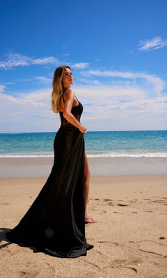 Wearing the Sheer-Waist Long Prom Dress: Nox Anabel E1711, a woman stands barefoot on a sandy beach near the ocean beneath a partly cloudy blue sky.