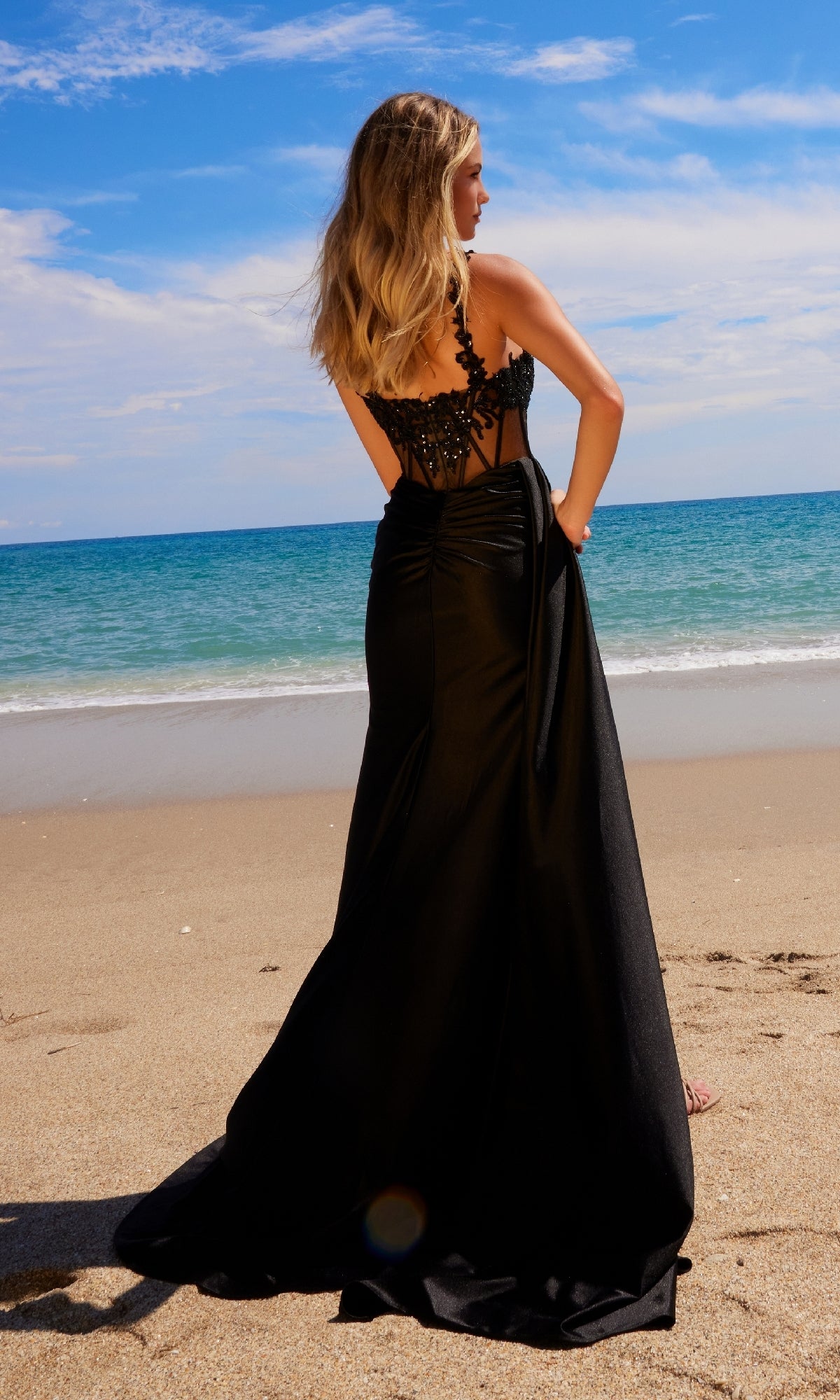 A woman in the Nox Anabel E1711 Sheer-Waist Long Prom Dress stands on a sandy beach, gazing at the ocean beneath a partly cloudy sky.