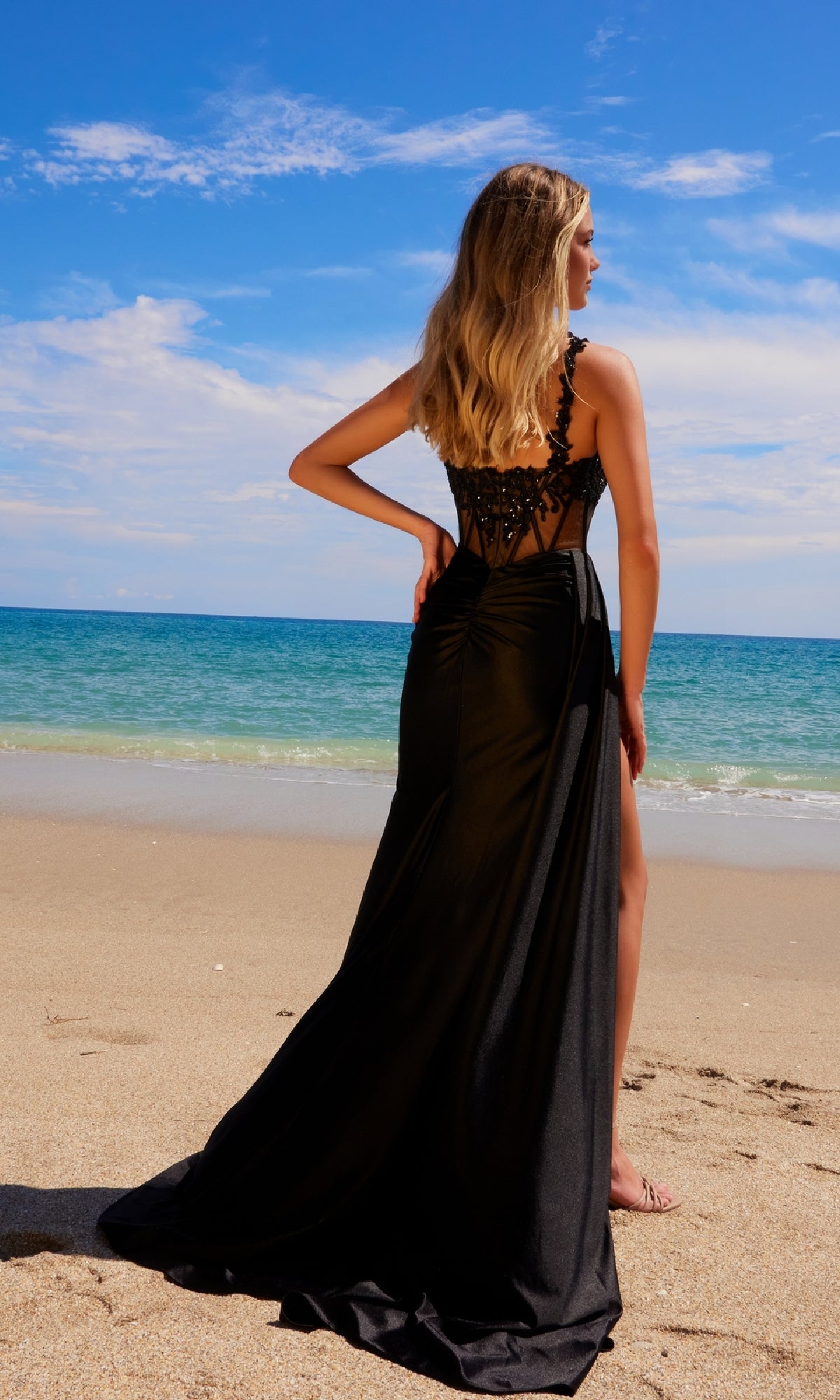 Wearing the Sheer-Waist Long Prom Dress: Nox Anabel E1711, a woman stands on a sandy beach facing the ocean beneath a partly cloudy sky.