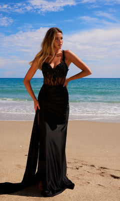A woman wearing the Sheer-Waist Long Prom Dress: Nox Anabel E1711 poses gracefully on a sandy beach, with the ocean and blue sky as her backdrop.