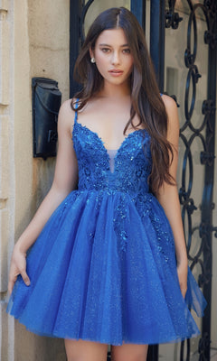 A woman stands before a wrought iron gate in the Nox Anabel E902 Glitter Tulle Homecoming Dress, a short blue design with sequin embroidery, floral embellishments, thin straps, and sparkling glitter tulle.