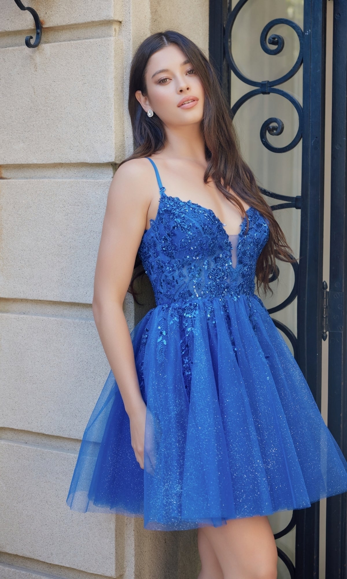 A woman stands before a stone wall and wrought iron gate, wearing the Nox Anabel E902 Glitter Tulle Homecoming Dress with sequin embroidery, a glitter tulle skirt, and delicate floral details.