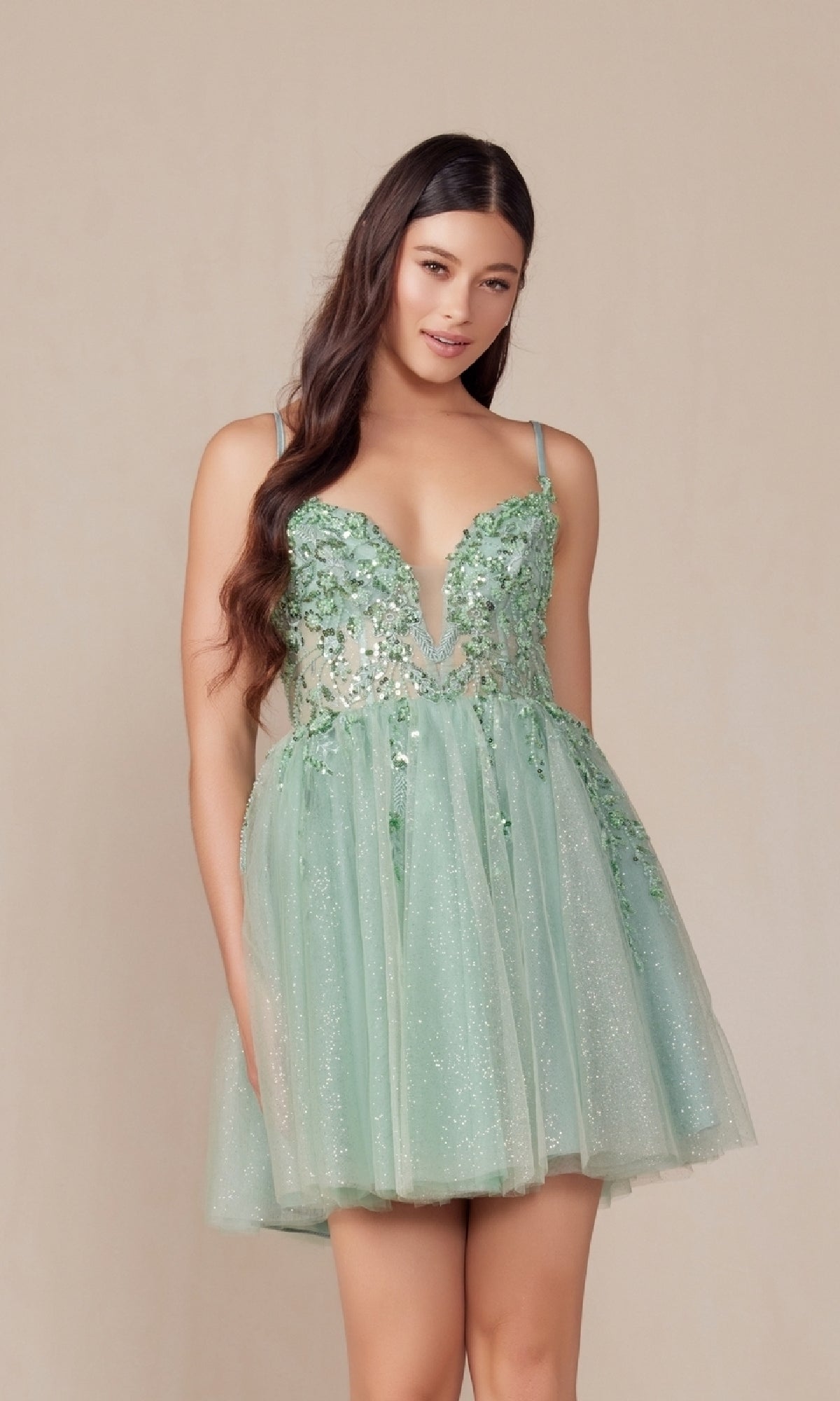 A woman wears the Nox Anabel E902 Glitter Tulle Homecoming Dress—a sleeveless, mint green gown with sequin embroidery, floral embellishments, a plunging neckline, and glitter tulle—standing against a plain beige background.