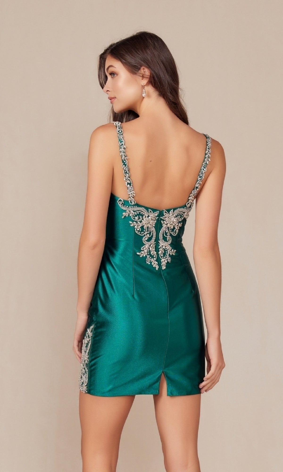 A woman models the Embellished Short Homecoming Dress E912, featuring green satin fabric, silver embroidery, an open back, and shoulder straps, against a plain beige background.