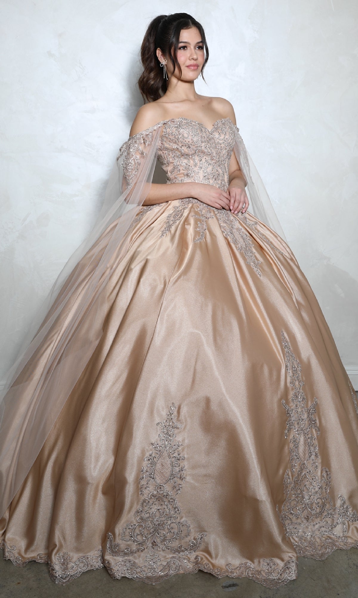 A young woman stands against a light wall wearing the Quinceanera Dress EK1002 By Eureka in champagne, featuring intricate lace details, a voluminous skirt, and an elegant lace-up back.