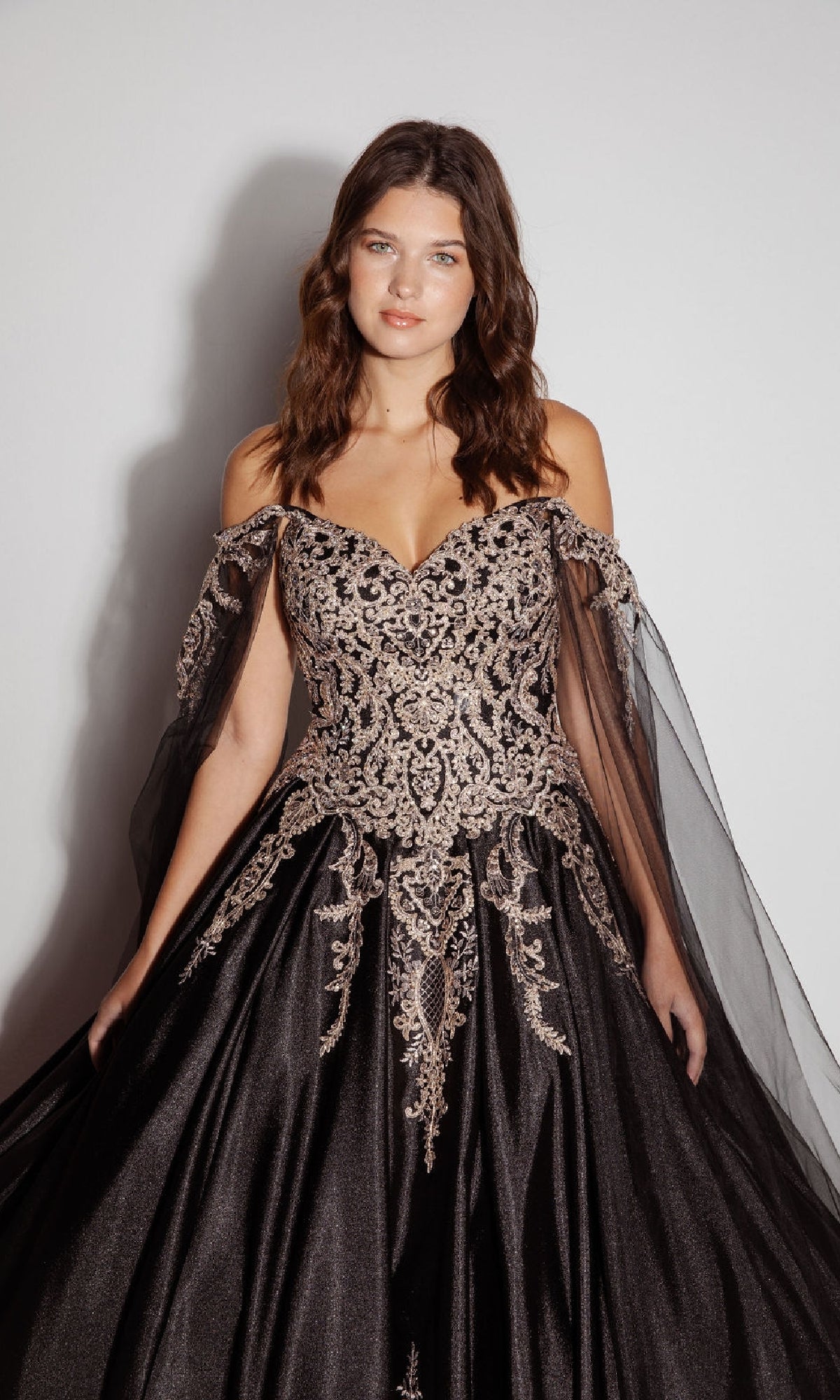 Woman modeling the Quinceanera Dress EK1002 By Eureka: an off-shoulder black ball gown with gold embroidery, sheer tulle sleeves, and a lace-up back, set against a plain light background.