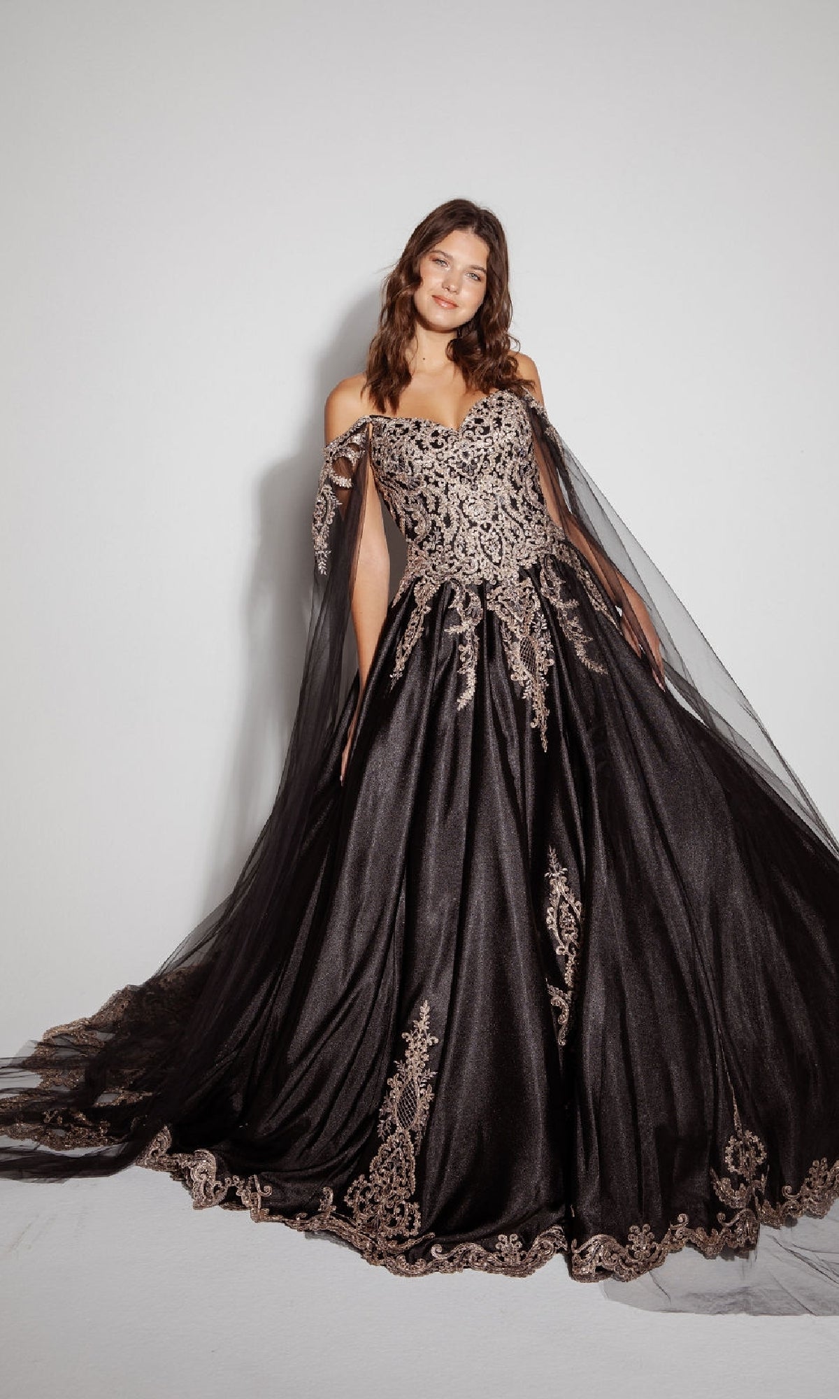 A woman wearing the Quinceanera Dress EK1002 By Eureka, an off-the-shoulder black ball gown with gold embroidery, poses against a plain white background, highlighting its classic elegance.