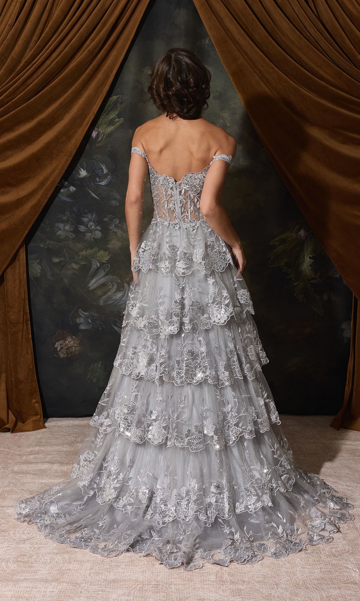 A person seen from behind wears an Off-Shoulder Long Ruffle Prom Dress: Eureka EK102, featuring an off-the-shoulder neckline and a multi-tiered ruffled skirt with silver floral embroidery.
