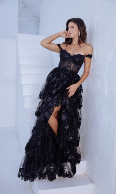 A woman poses on white stairs against a textured wall, wearing the Off-Shoulder Long Ruffle Prom Dress: Eureka EK102, one hand touching her face and the other elegantly holding her gown.