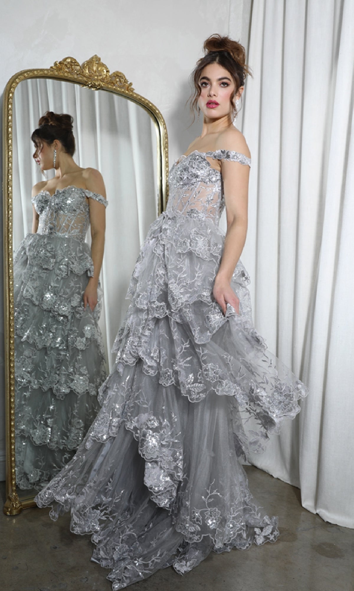 A woman in the Off-Shoulder Long Ruffle Prom Dress: Eureka EK102, featuring layered silver lace, stands before a gold-framed mirror with white curtains behind her.