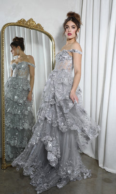 A woman in the Off-Shoulder Long Ruffle Prom Dress: Eureka EK102, featuring layered silver lace, stands before a gold-framed mirror with white curtains behind her.