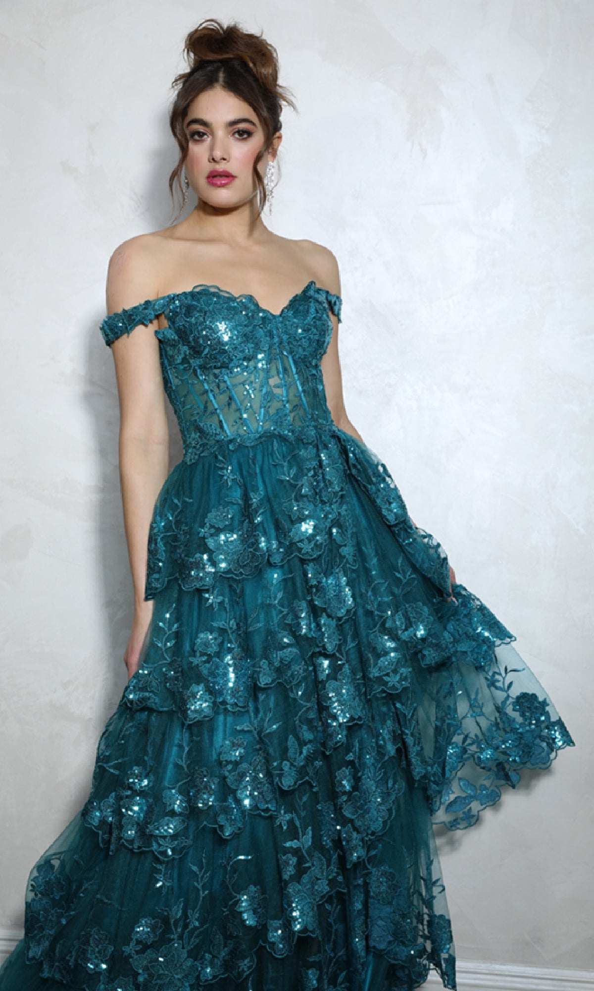 A woman stands against a light textured wall in the Eureka EK102 Off-Shoulder Long Ruffle Prom Dress—teal with layered ruffles and floral sequin embellishments—an eye-catching formal gown perfect for special occasions.