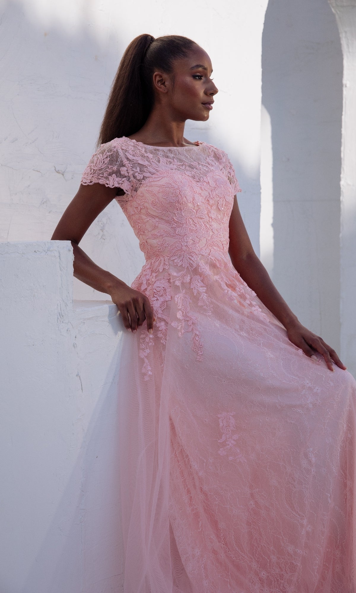 A woman in the Modest Short-Sleeve Prom Gown: Eureka EK104 stands against a white textured wall and archway, looking to the side.