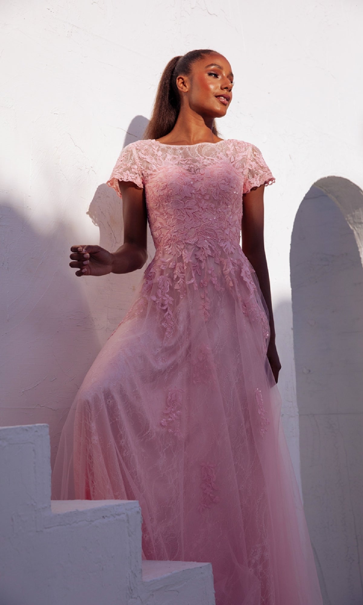 A woman in the Modest Short-Sleeve Prom Gown: Eureka EK104 stands by a white textured wall with arched openings, gazing upward and resting her hand on a stair railing, her pink floral-lace evening dress adding elegance to the scene.